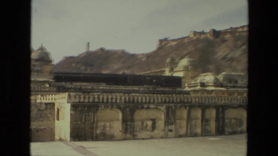 1974 India 8mm frame showing a long weathered palace facade, arched openings, small domes, and fort-like walls along the hillside.