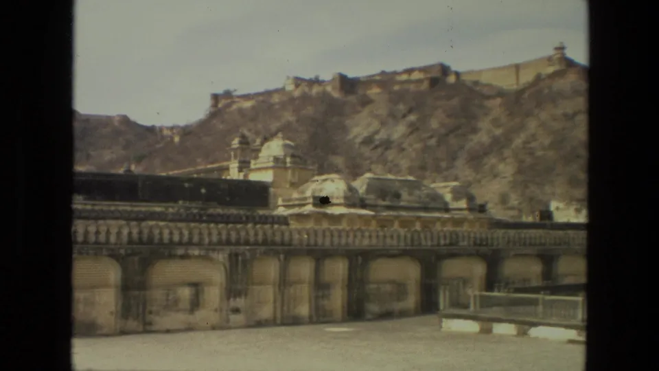 1974 India home-movie frame with an extended palace wall, domed roofline details, hillside fortifications, and an open paved foreground.