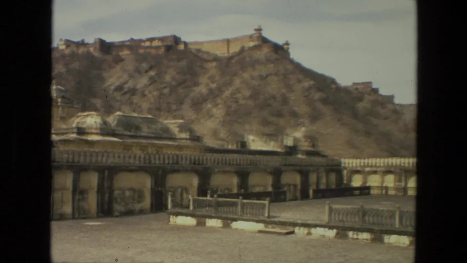 1974 India 8mm frame with a palace-like arched courtyard, domed masonry roof, and a rocky hillside rising to fort-like walls.