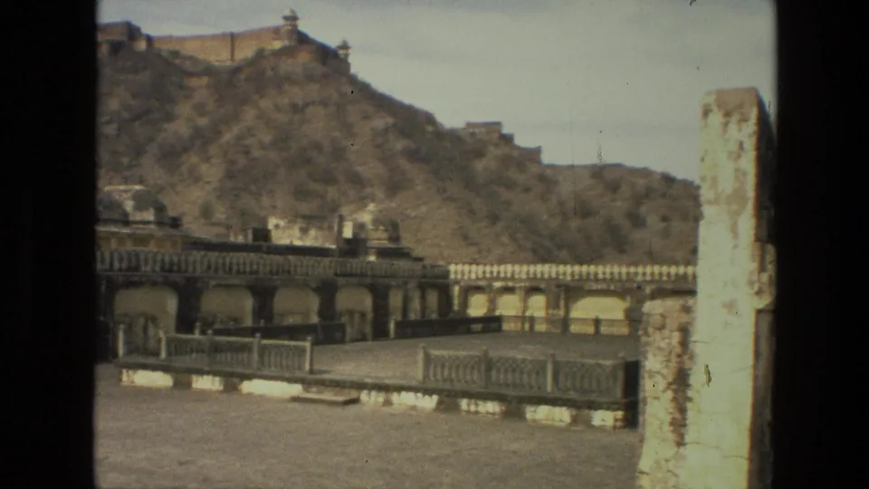1974 India 8mm frame showing a weathered palace courtyard, balustrade, arched walls, dry hillside, and a pale masonry parapet at right.