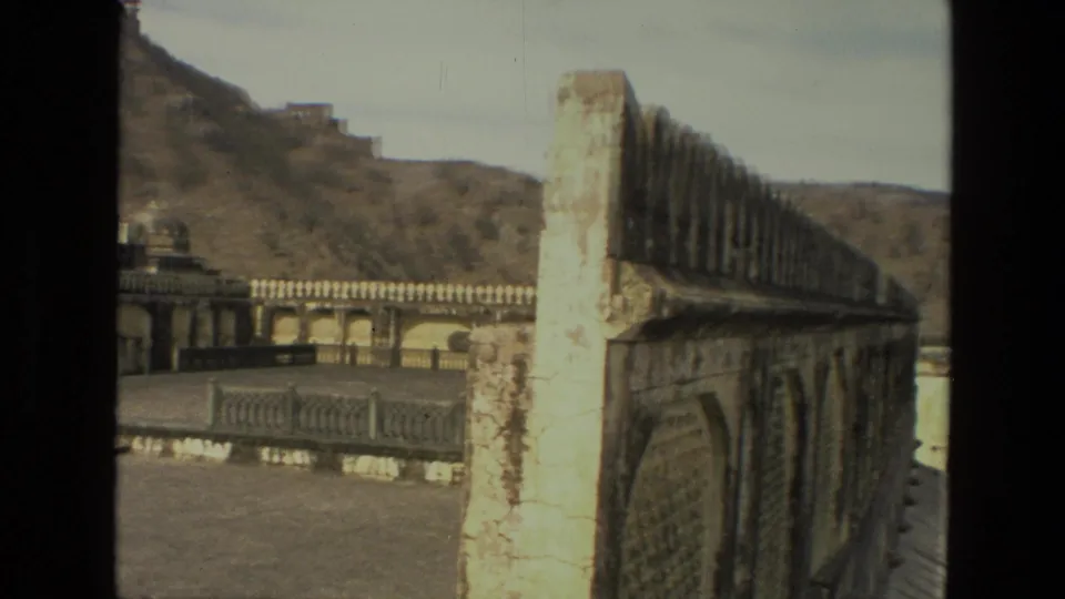 1974 India 8mm frame showing a long weathered masonry wall, courtyard balustrade, arched building edges, and hillside ruins in the distance.