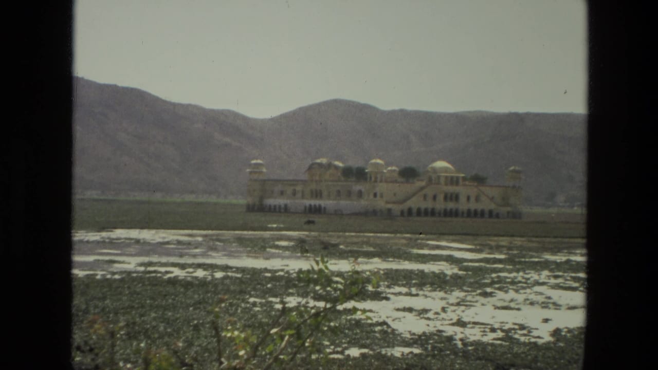 1974 India 8mm frame showing a distant domed palace-like building across aquatic plants and shallow water, with hazy mountains beyond.