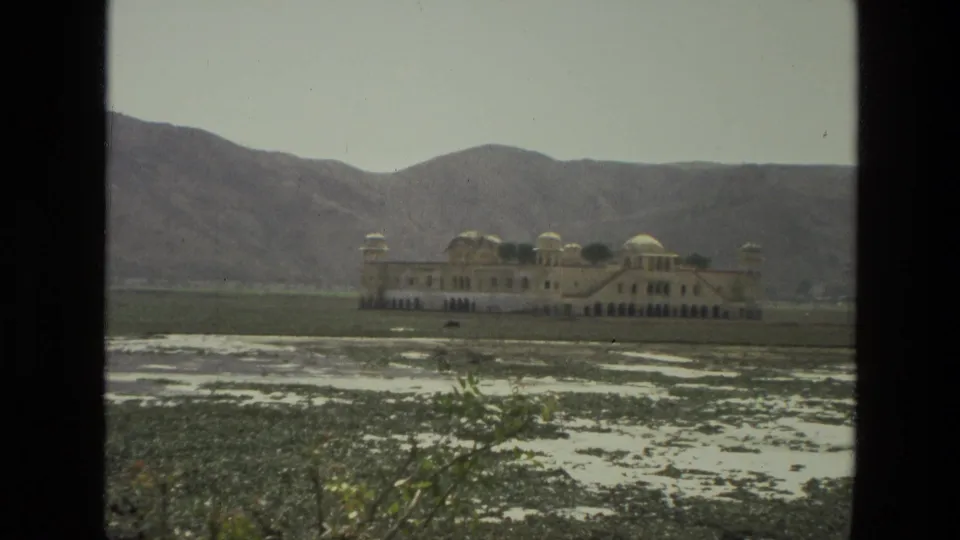 1974 India 8mm frame showing a distant domed palace-like building across aquatic plants and shallow water, with hazy mountains beyond.