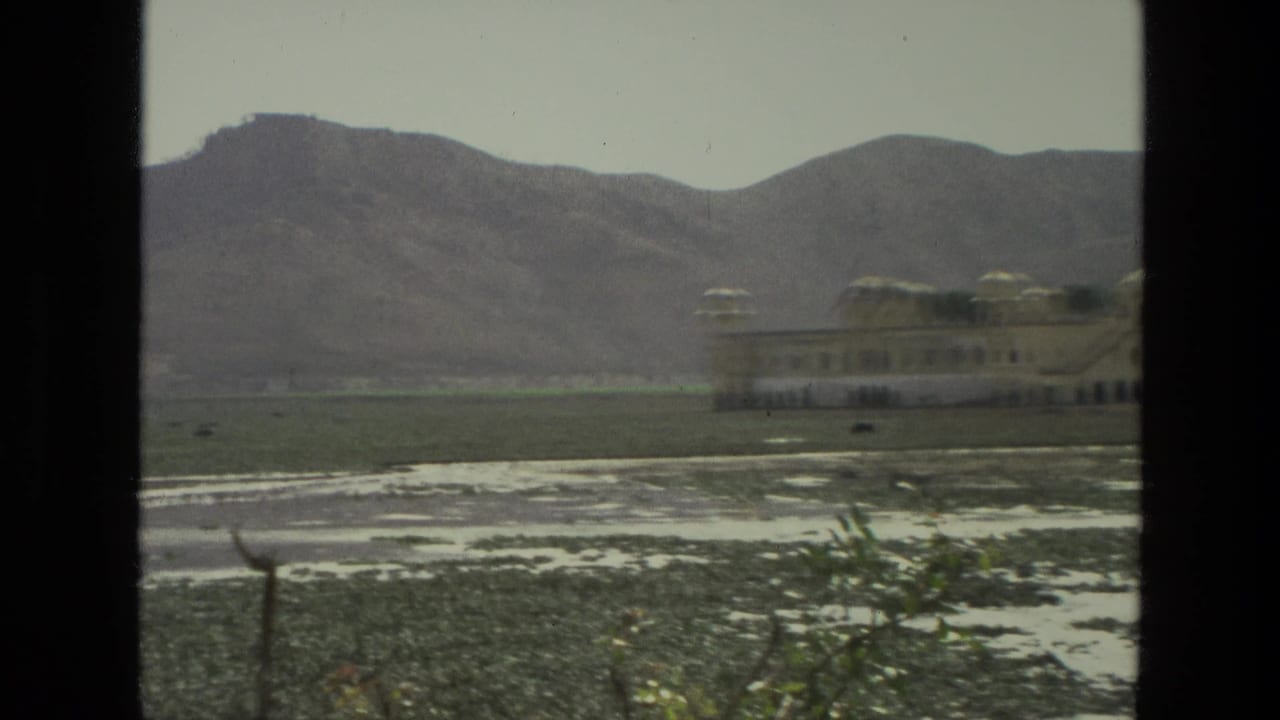 1974 India 8mm frame with a palace-like domed structure partly at right, seen across a flat wetland with mountain ridges behind.