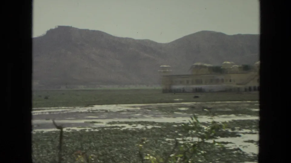 1974 India 8mm frame with a palace-like domed structure partly at right, seen across a flat wetland with mountain ridges behind.