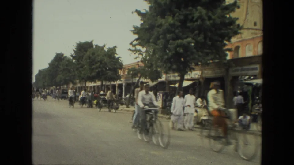 1974 India 8mm street scene with cyclists, pedestrians, trees, shopfronts, and roadside activity.