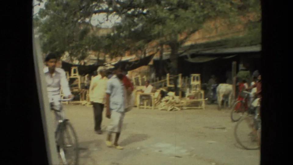 1974 India 8mm frame showing men walking and cycling past roadside shops, seating, and trees.