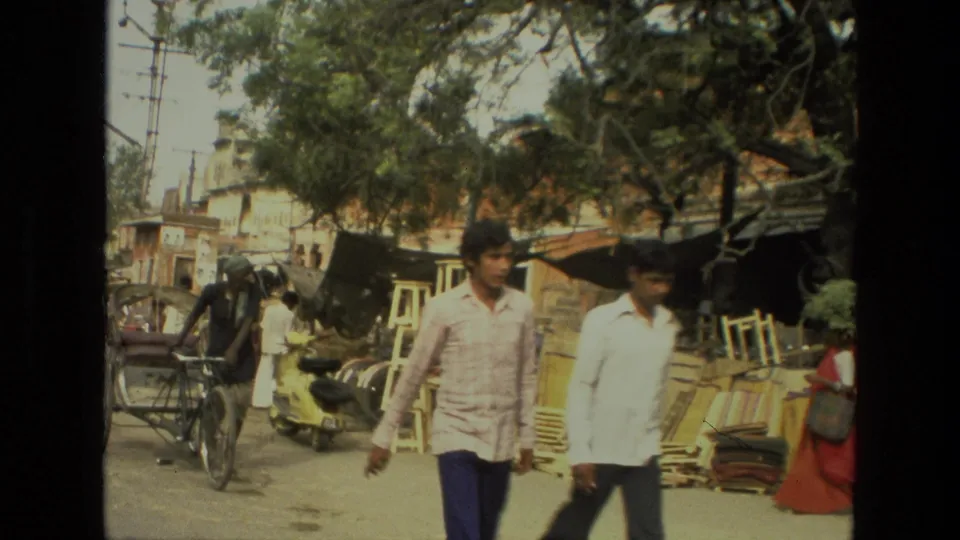 Two men walk through a 1974 India market street as a horse-drawn cart and roadside wooden furniture stalls sit behind them.