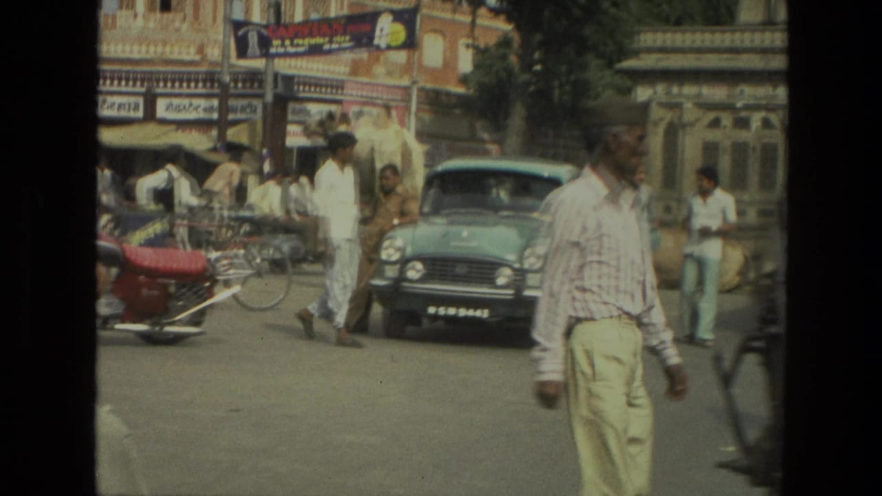 Busy Indian street with pedestrians, cycle rickshaw, older car, storefront signs, and ornate buildings.