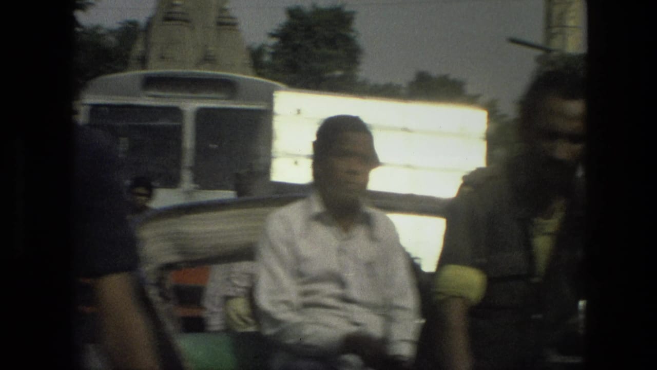 Man seated in a rickshaw or small vehicle with a bus-like vehicle and bright side panel behind him.