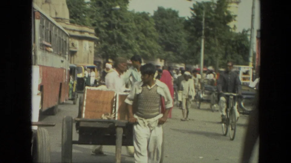 1974 India street scene with a bus or tram-like vehicle at left, a handcart in front, pedestrians, bicycles, and rickshaws in traffic.