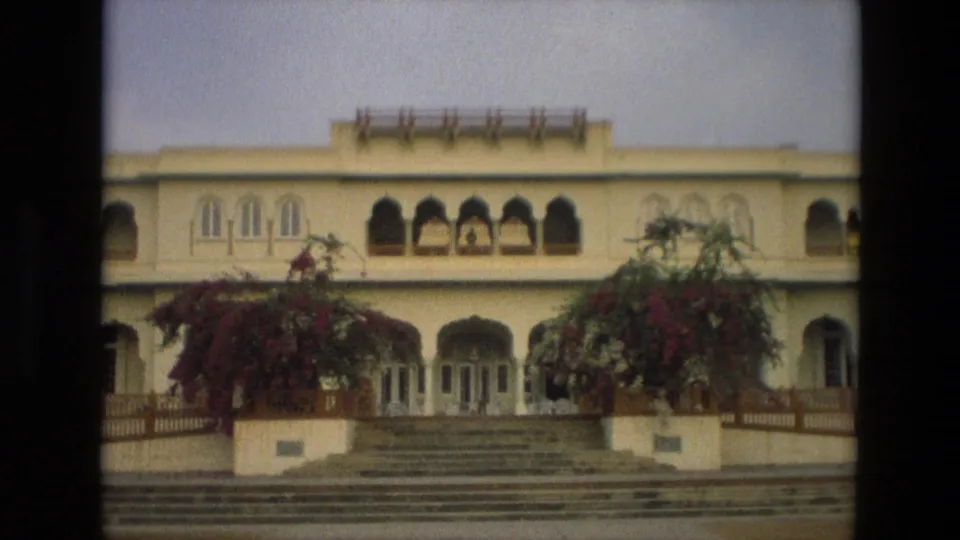 Light-colored Indian building with arched windows, entrance steps, railings, and flowering vines, seen in soft 8mm color.