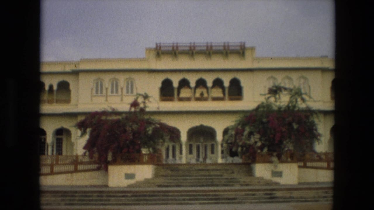 1974 India home-movie frame showing a cream palace-like facade with arched windows, broad steps, railings, and bougainvillea.