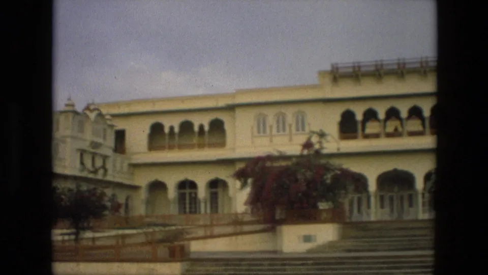 1974 India frame of the palace exterior during a pan, showing arched openings, cream walls, steps, and flowering plants.