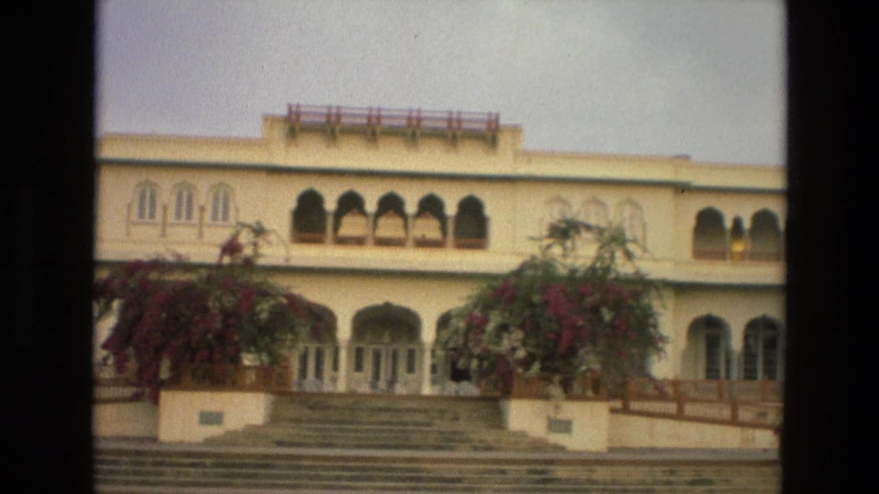 1974 India 8mm frame showing a cream palace or heritage building facade with arched openings, steps, railings, and flowering shrubs.