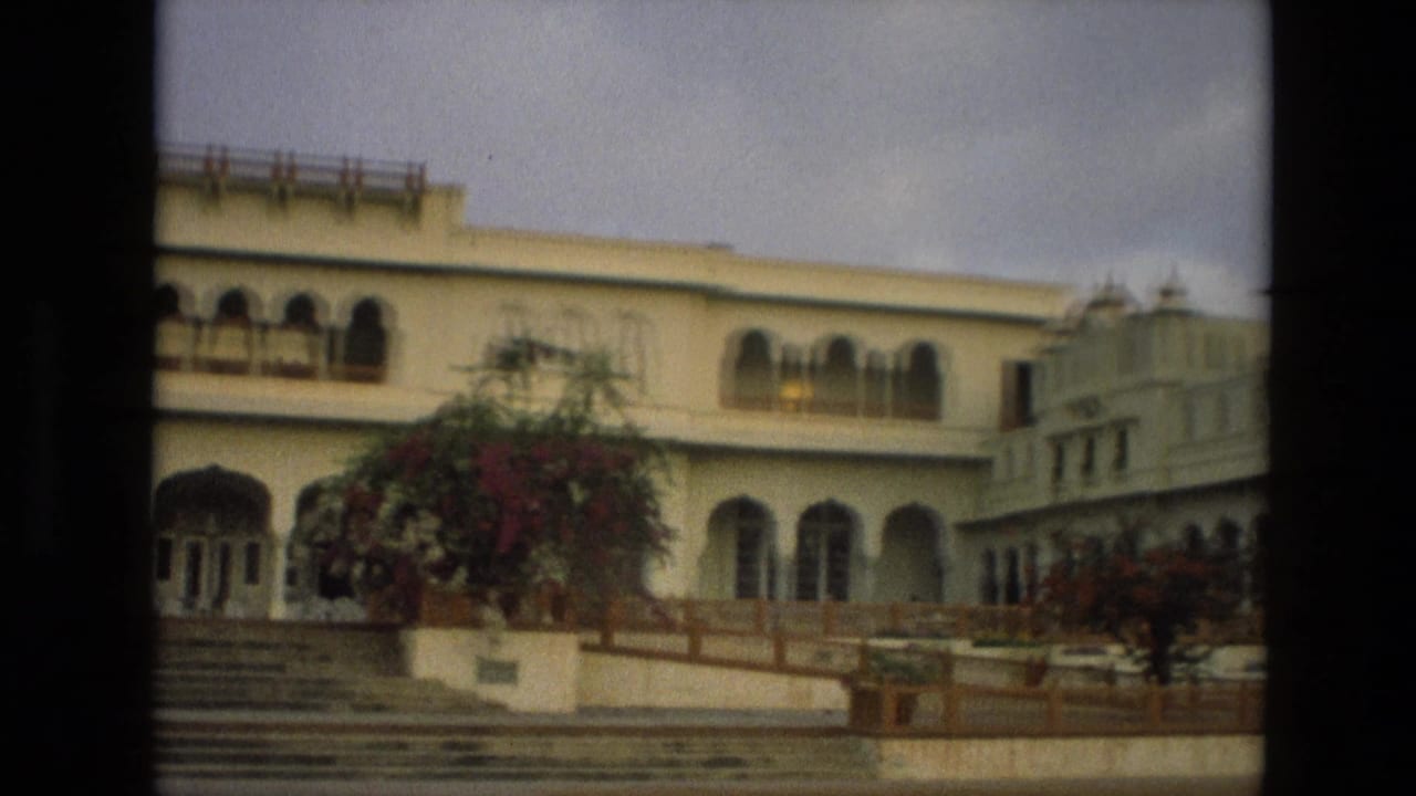 1974 India 8mm frame of a palace-like exterior with arched colonnades, balcony details, terrace railings, and flowering landscaping.