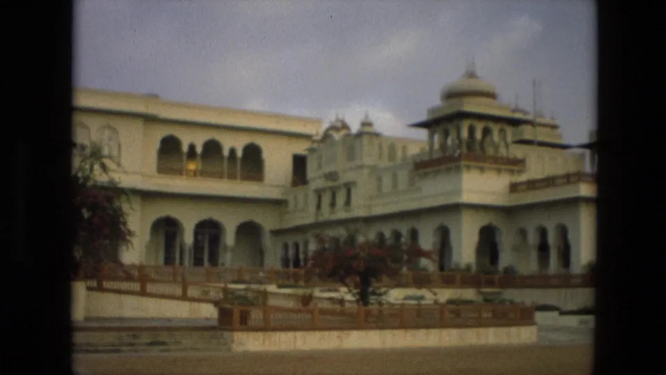 1974 India 8mm frame showing the building from an angle, with a domed chhatri, arched openings, terrace railing, and landscaped shrubs.