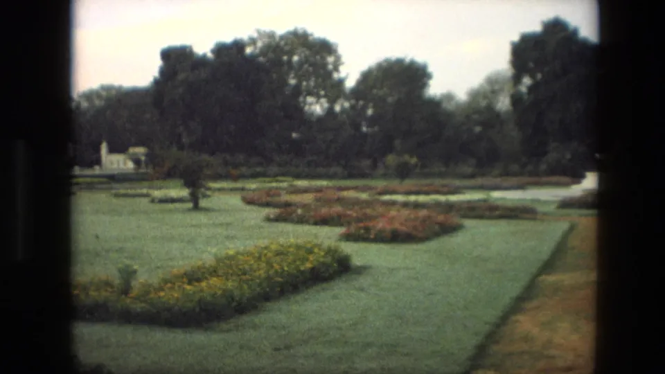 1974 India 8mm view across landscaped palace gardens with green lawns, rounded flowerbeds, path edge, trees, and a distant white garden structure.