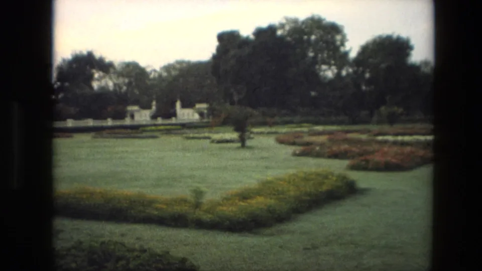 1974 India 8mm frame showing formal garden lawns, colorful flowerbeds, distant white structures, and mature trees under soft exposure.