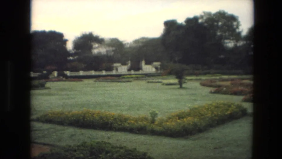 1974 India 8mm frame showing a formal garden with open lawn, low flowerbeds, a pale boundary wall, and mature trees.