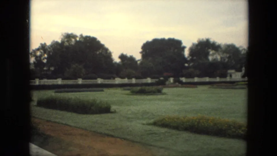 1974 India 8mm still of a quiet formal garden, with clipped grass, planted beds, a pale wall, and mature trees beyond it.