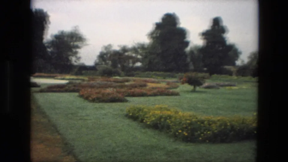 1974 India home-movie frame showing a garden path, manicured lawns, colorful flowerbeds, and trees in an overcast landscape.