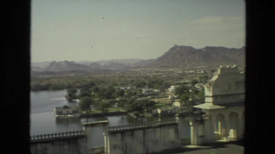 1974 India home-movie view from above, with water at left, a domed pavilion at right, trees, low buildings, and mountains.