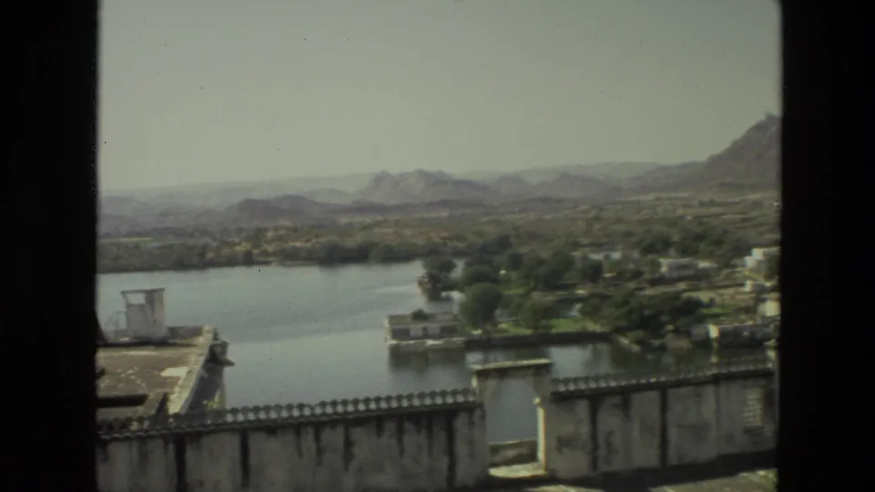 1974 India frame showing a lake or pond below a terrace wall, with greenery, scattered buildings, and rocky hills in the distance.