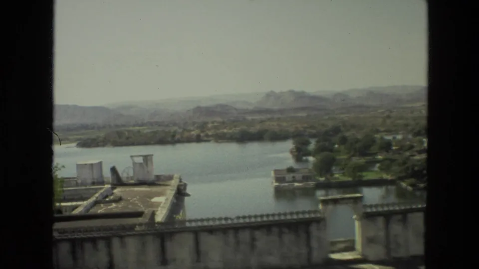 1974 India 8mm frame showing a lake from above, with a weathered parapet, low rooftops, trees, and distant hills under haze.