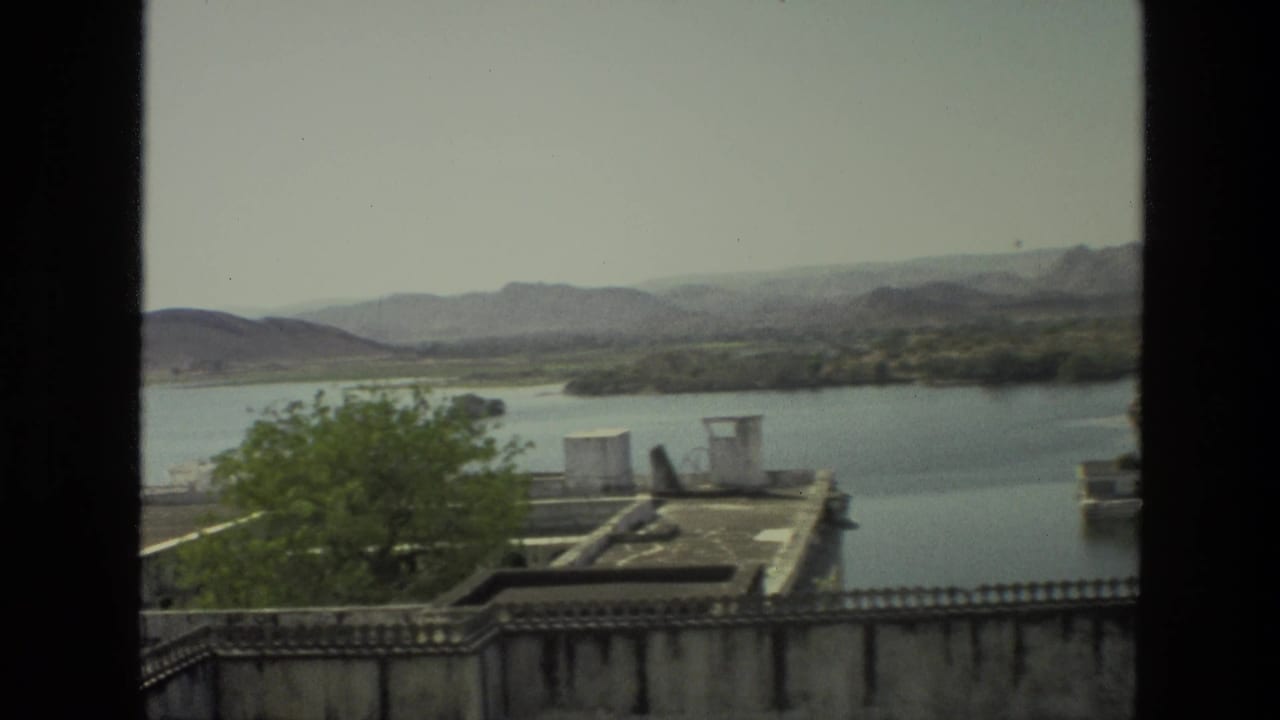 Lake and hills seen past a leafy tree and rooftop area, with a dark vertical film edge at left.