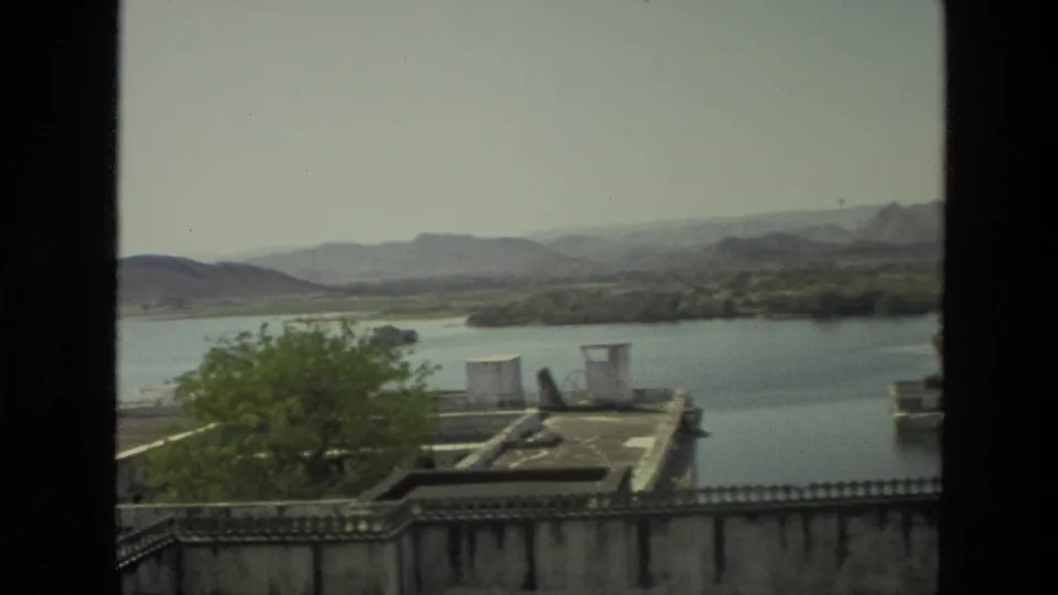 1974 India travel footage frame with lake water, distant hills, a leafy tree, and flat rooftop structures in soft 8mm color.