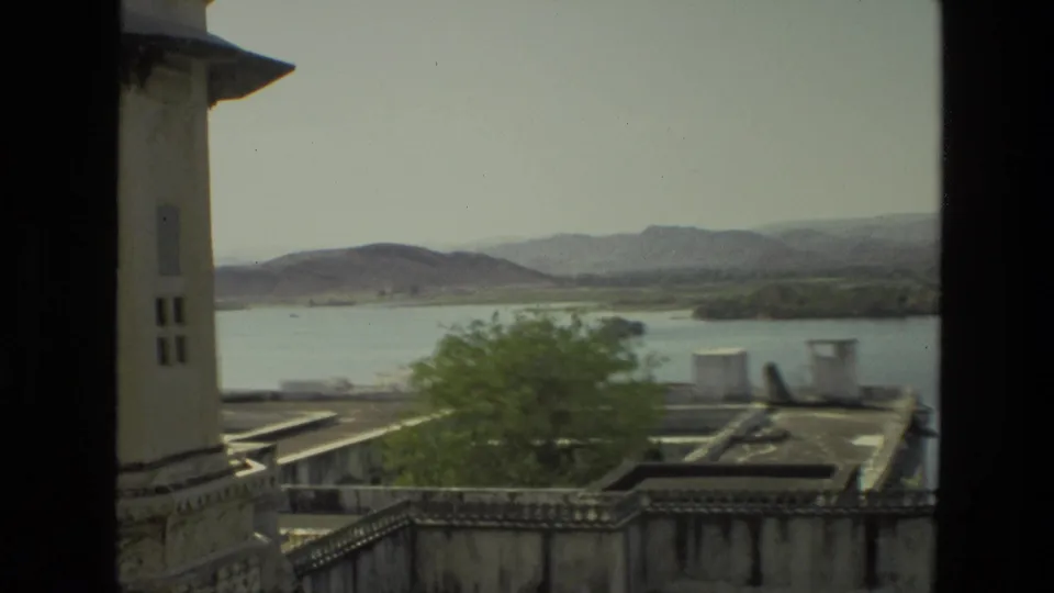 1974 India 8mm frame looking past a building edge and stone parapet toward a lake, trees, rooftops, and hazy hills.