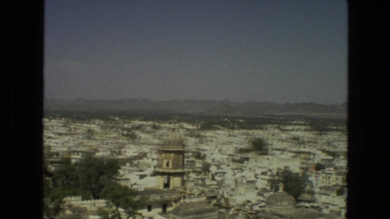 1974 India frame showing a broad cityscape of pale buildings, trees, distant hills, and a small dark figure or animal on a rooftop.