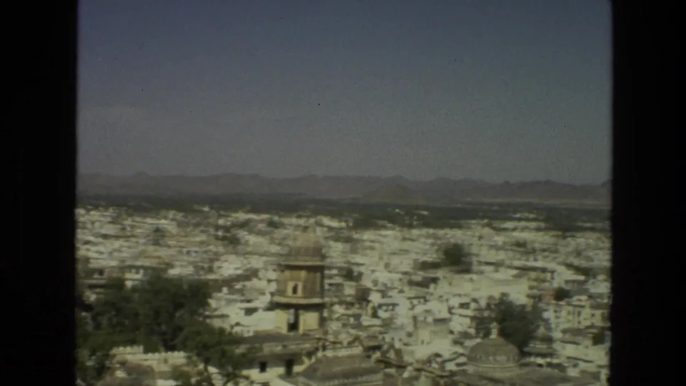1974 India frame showing a broad cityscape of pale buildings, trees, distant hills, and a small dark figure or animal on a rooftop.