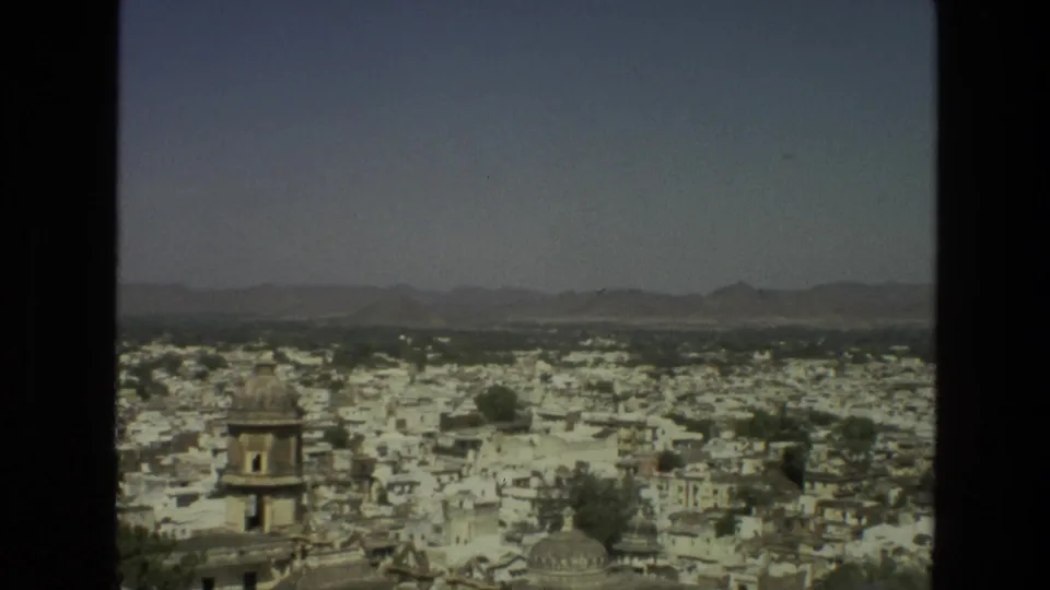 1974 India home-movie frame showing whitewashed rooftops, a small tower, trees, and hazy hills from an elevated viewpoint.