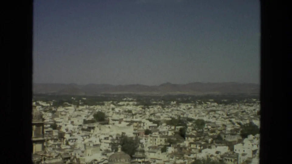 1974 India frame showing a dense city panorama with a domed roof in the foreground, trees, low hills, and bright sky.
