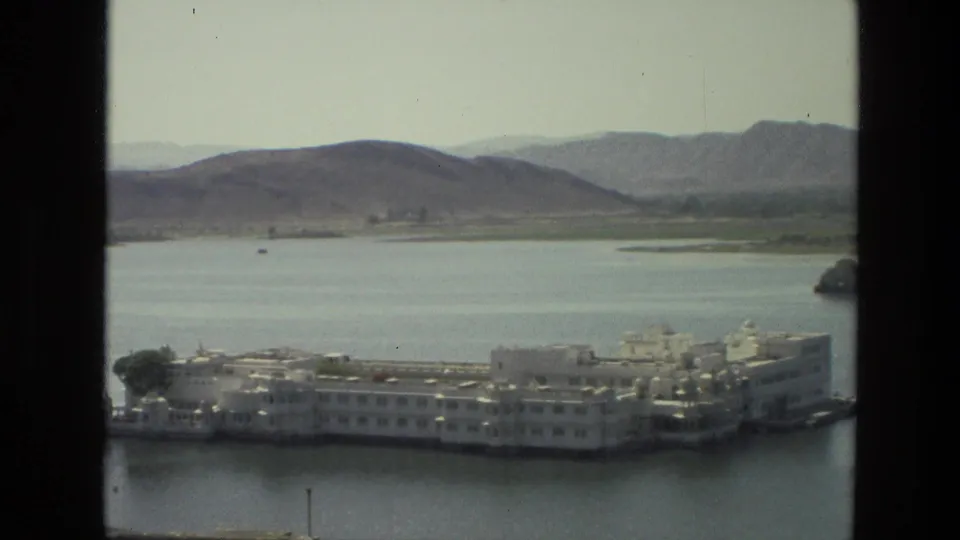 1974 India 8mm frame showing a white palace-like complex on a calm lake, backed by hazy hills and shoreline vegetation.