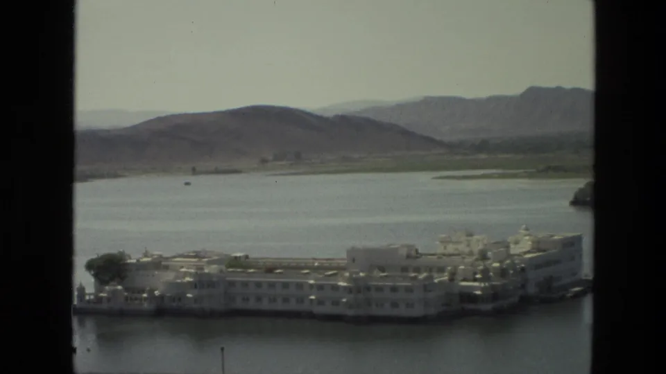 1974 India 8mm frame showing a white palace-like island complex on a lake, with hazy hills and dark window framing.