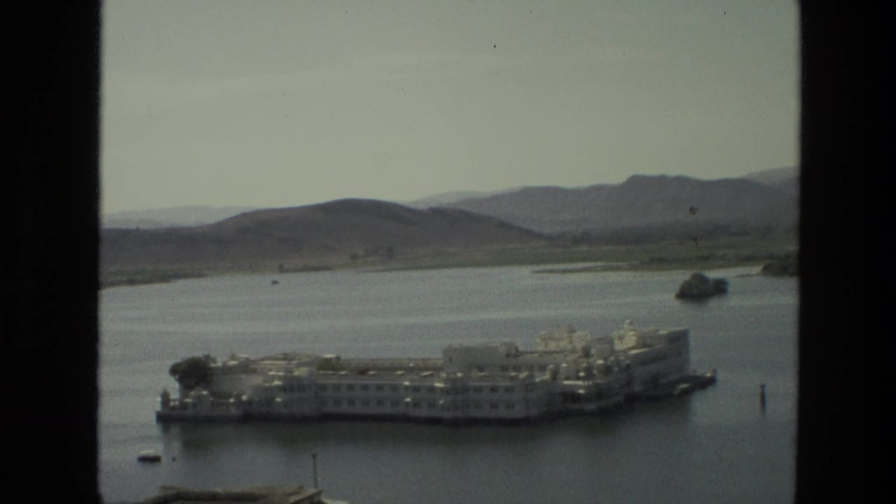 1974 India travel frame with a white lakeside architectural complex, small boats, distant hills, and dark window edges.