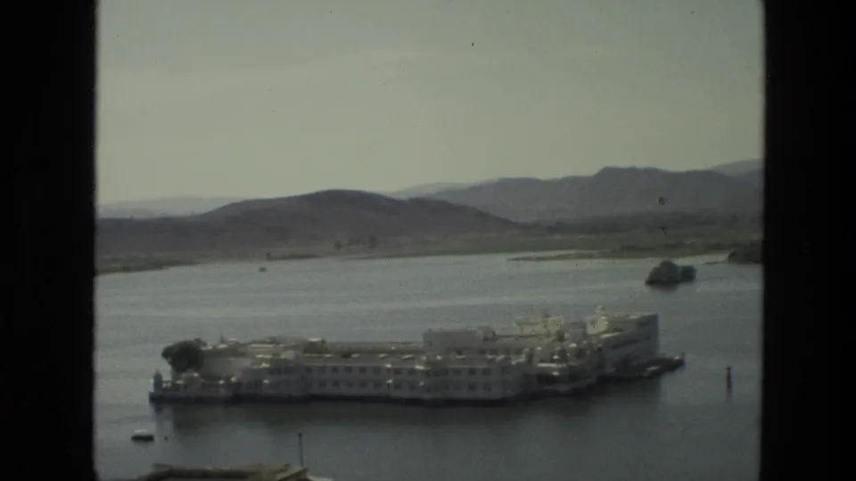 1974 India travel frame with a white lakeside architectural complex, small boats, distant hills, and dark window edges.