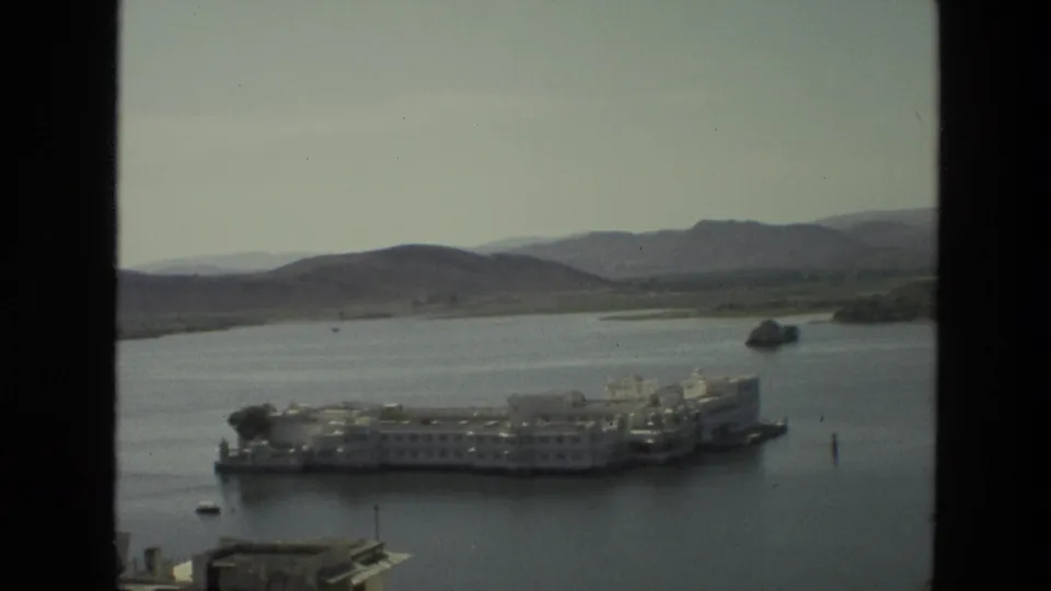 Wider 1974 India 8mm view of a white island complex on calm water, backed by arid hills and a hazy sky.
