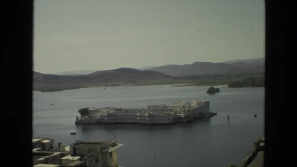 Wide 1974 India home-movie frame showing white island buildings on a lake with hills, foreground rooftops, and window-like borders.