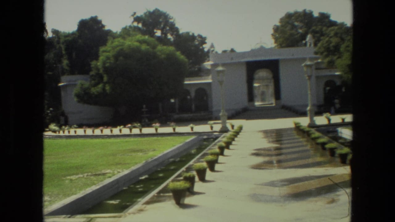 India 1974 garden scene with a pale arched gateway, flanking lawns, potted plants, trees, and dark vertical edges from the filming position.