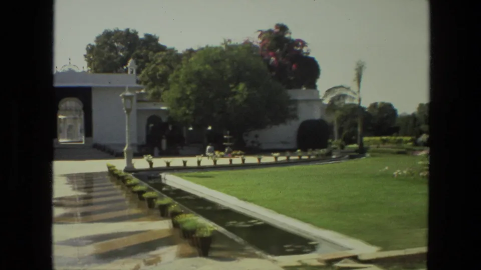 India 1974 home-movie frame showing a landscaped garden, white arched structure, lawns, water channel, palms, and distant visitors near the path.