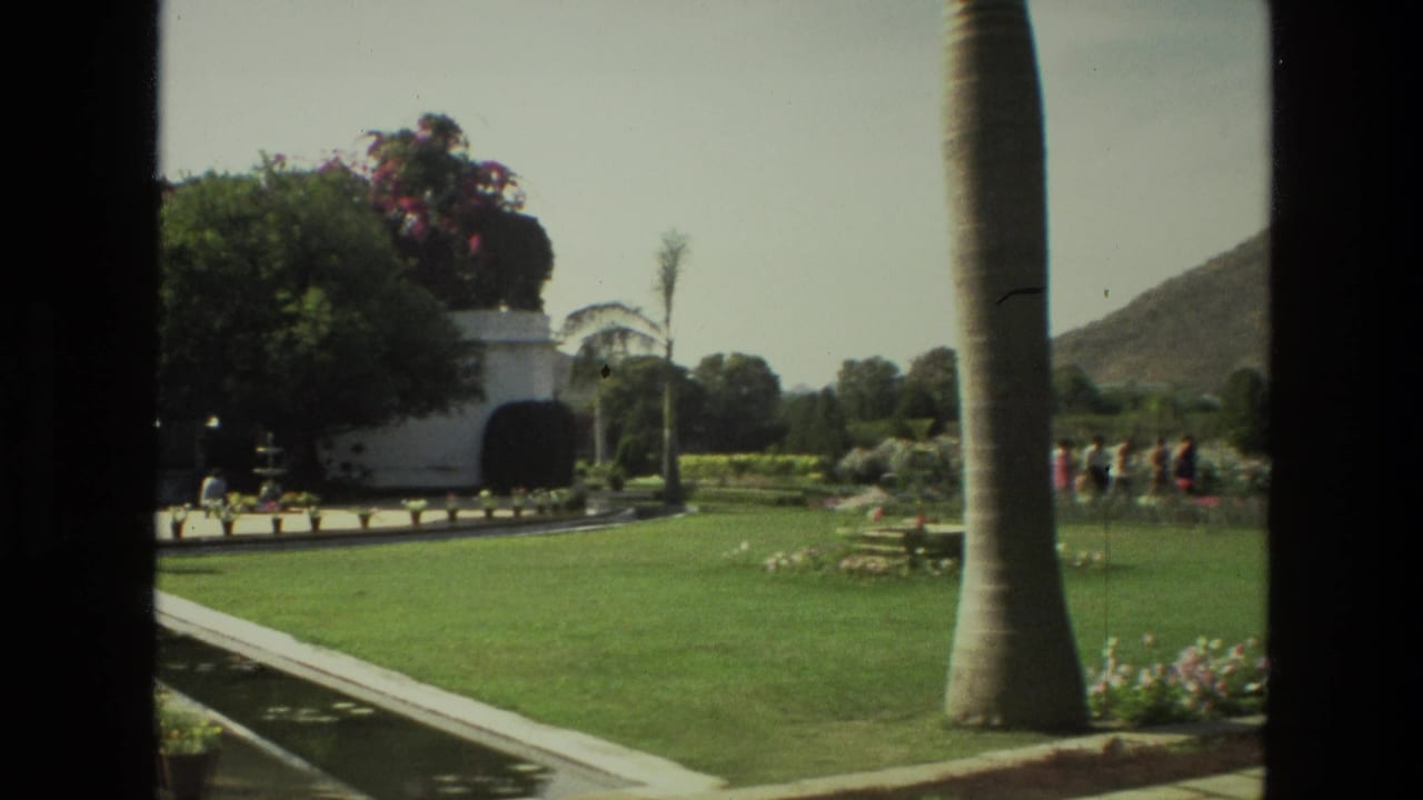 1974 India home-movie frame with palm trunk in the foreground, formal lawn, flower beds, white garden structure, and small distant visitors.