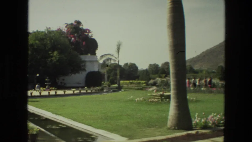 1974 India home-movie frame with palm trunk in the foreground, formal lawn, flower beds, white garden structure, and small distant visitors.