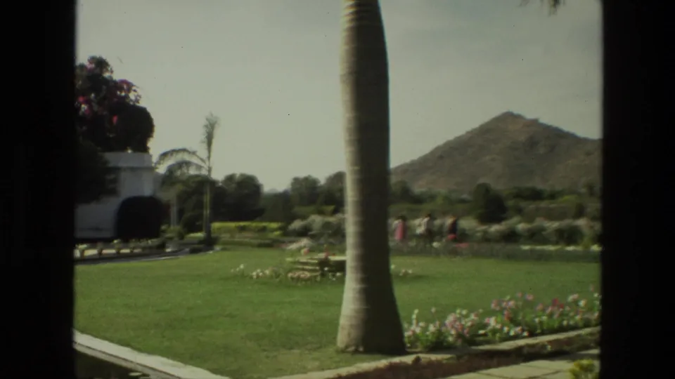 1974 India 8mm frame showing a landscaped lawn, foreground palm trunk, hillside, flower beds, and visitors walking through the grounds.