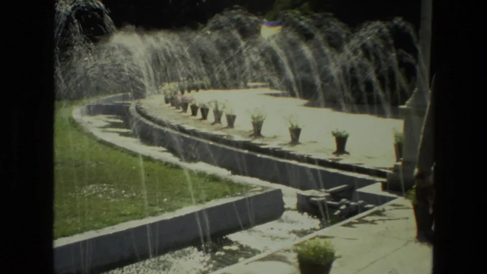 1974 India home-movie frame of a curved fountain with arcing water jets, potted plants, lawn edge, and walkway in a formal garden.