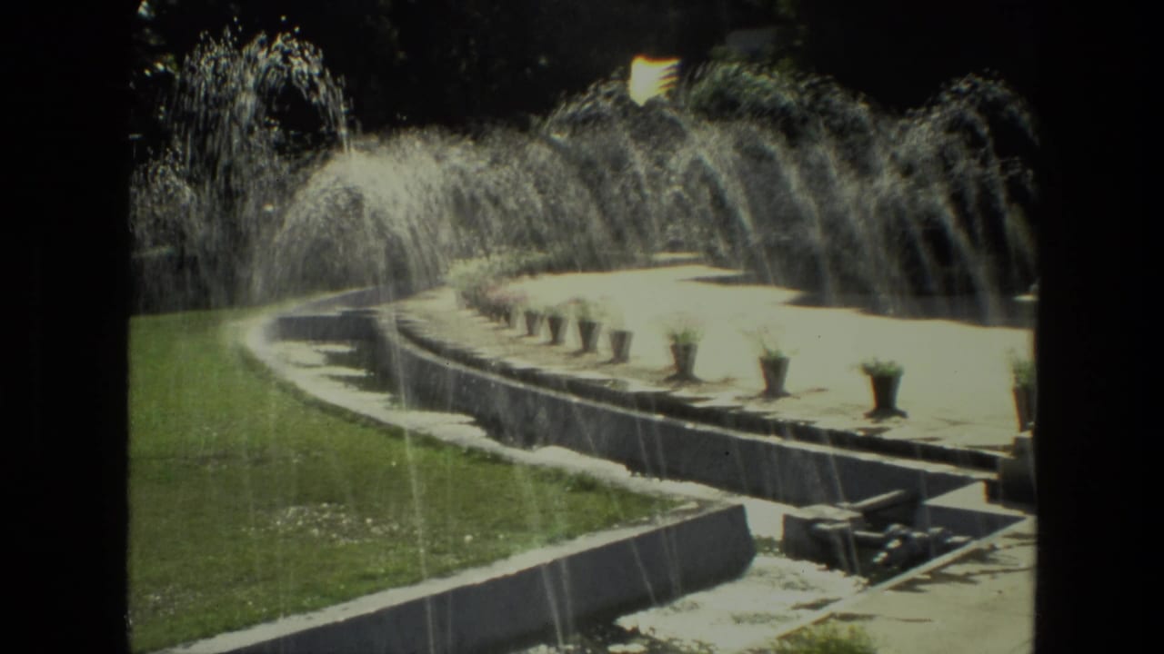 Curved fountain basin with arcing water jets beside a lawn and terrace planters.