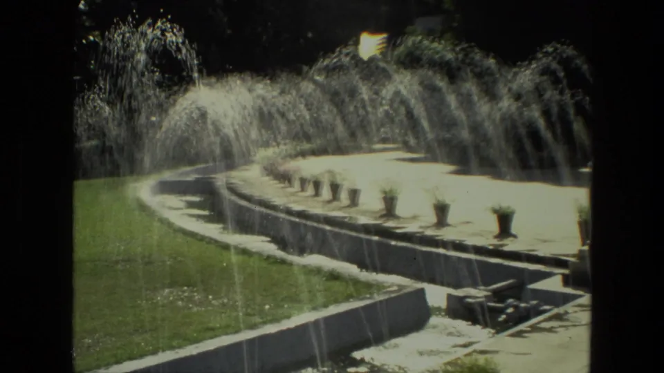 1974 8mm home-movie view in India of a curved garden fountain, lawn, stone edging, and potted terrace plants in bright sun.